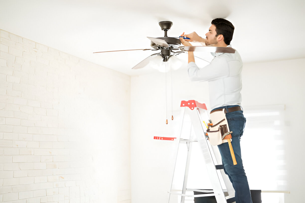 A man on a ladder in a bright room uses a screwdriver to fix an Australian ceiling fan, wearing a tool belt.