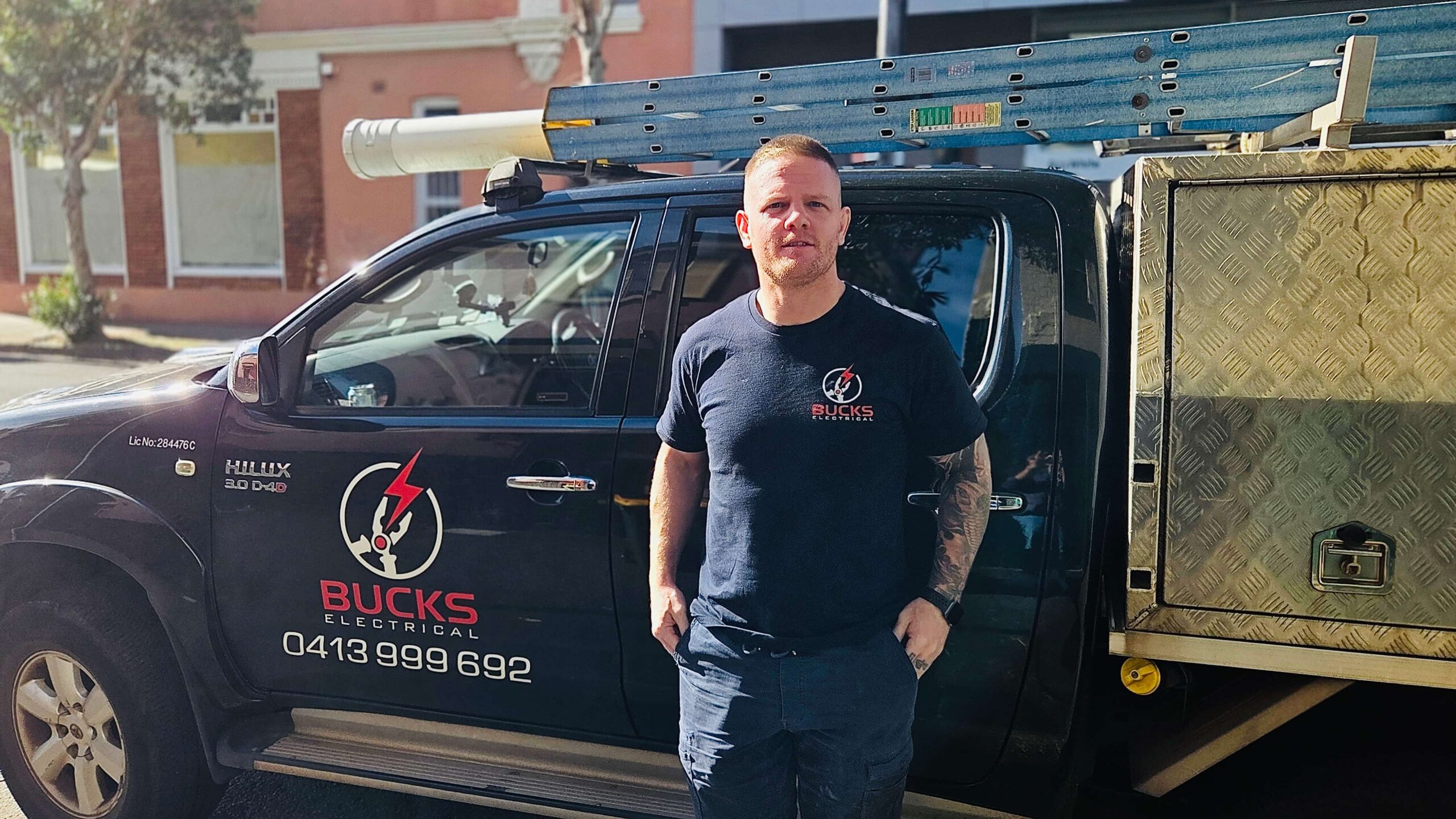 Stephen Buckle stands beside her Bucks Electrical truck with ladder and tool storage on a sunny day.