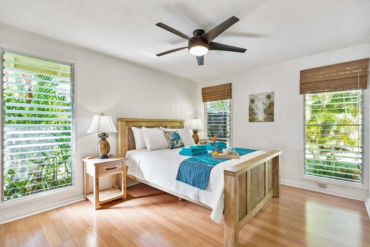 Bright bedroom with wooden bed, ceiling fan, tropical decor, bedside lamps, and folded towels on the bed.