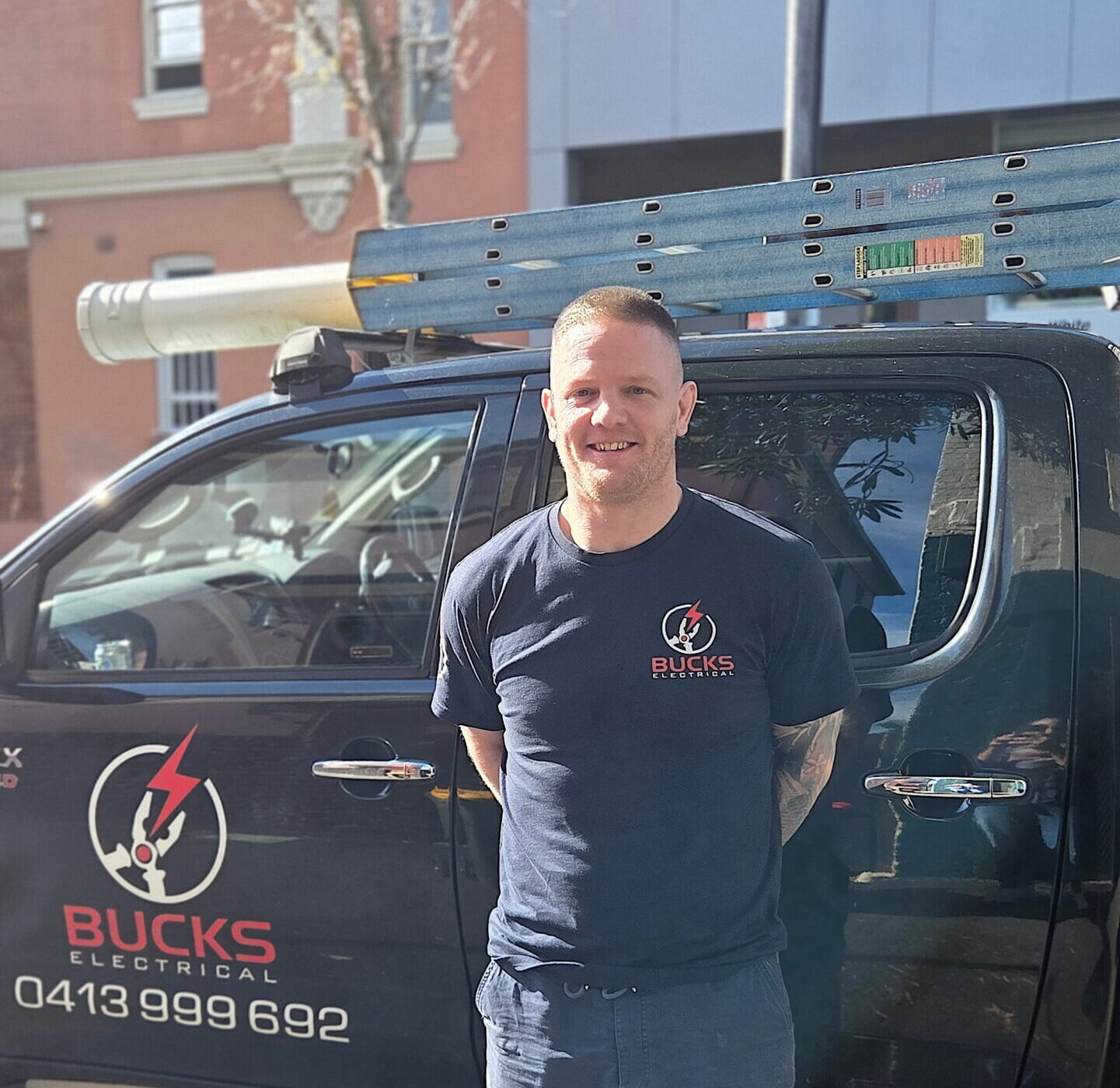 Stephen Buckle, a Bucks Electrical technician, stands by a branded truck with ladder on a city street.