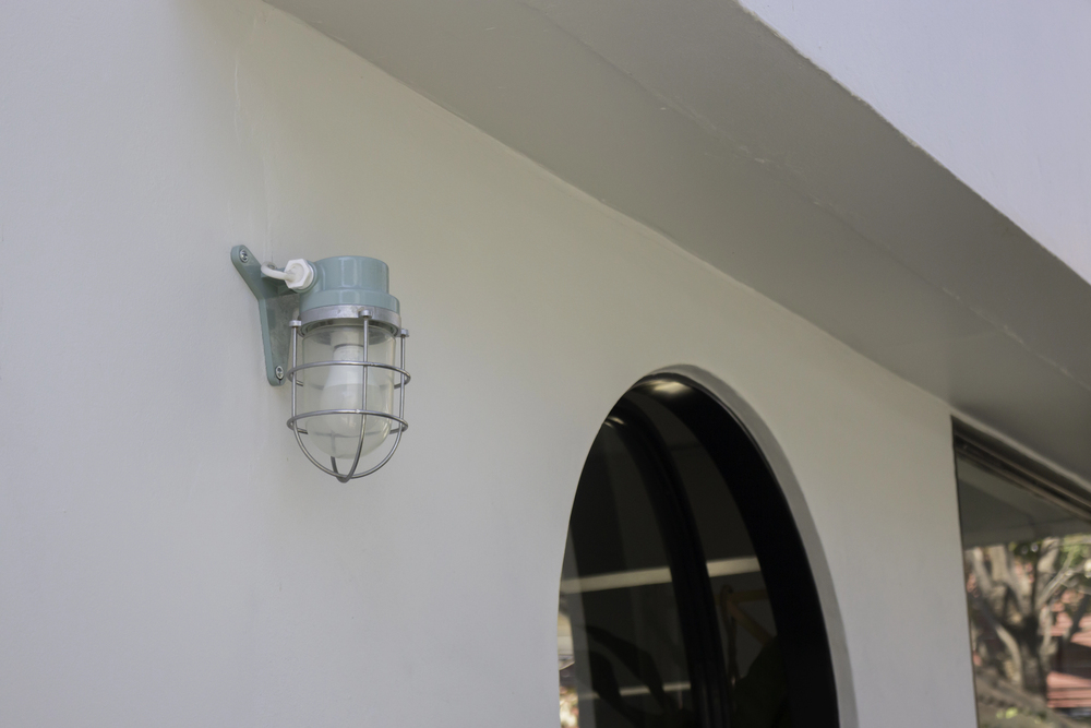 Outdoor wall-mounted light with cage near black arched window on white building, surface downlight style.