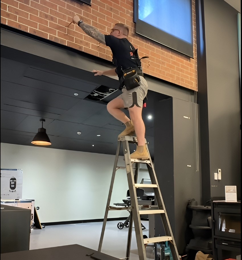 Stephen from Bucks Electrical, in workwear, on ladder reaching a high window; tools nearby, her name Stephen.